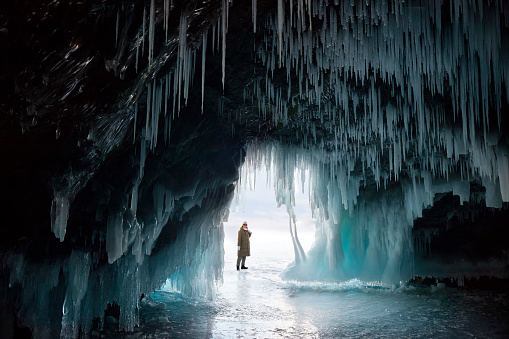 View from an ice cave or grotto with huge blue icicles. A woman admires the winter landscape.