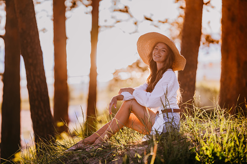 Young female swedish traveler woman in straw hat, white dress with buttons, looking at camera touching hat sitting at wild forest. Attractive blonde caucasian girl traveling. Leisure, people at nature