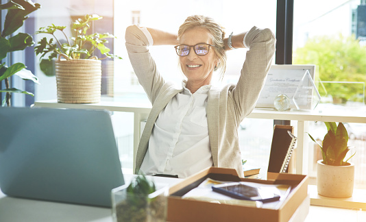 Happy mature manager satisfied and relieved to be done with deadlines and tasks. Business woman feeling accomplished and enjoying a relaxing break to stretch with hands behind her head in an office.