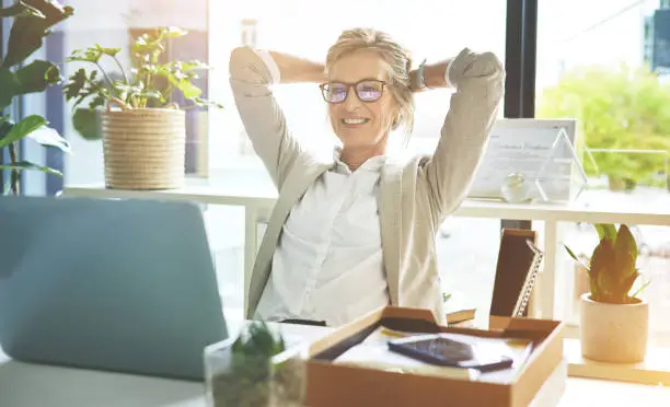 Photo of Happy mature manager satisfied and relieved to be done with deadlines and tasks. Business woman feeling accomplished and enjoying a relaxing break to stretch with hands behind her head in an office.