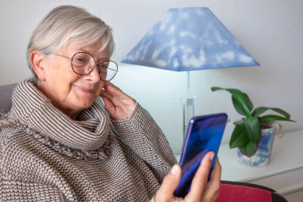 テクノロジーとソーシャルメディアを楽しむ携帯電話を使って自宅に座っている美しいシニア女性の肖像画 - 古い世代と新しいテクノロジーのコンセプト - old armchair women senior adult ストックフォトと画像