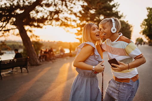 Two gay female friends listen to music while sharing wireless headphones. Two young beautiful girls are walking through the city and listen to music