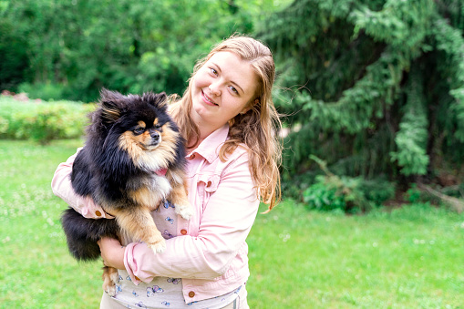 Portrait of young blond cheerful caucasian woman in casual pink clothes holding on hands and hugging her fluffy black dog pet Spitz Pomeranian, in summer park or backyard on green grass loan