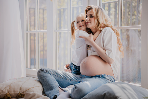 Pretty pregnant swedish woman with daughter in white t-shirt and blue jeans sitting on the floor with pillows hugging daughter tenderly, feeling love.
Maternity and pregnancy concept.