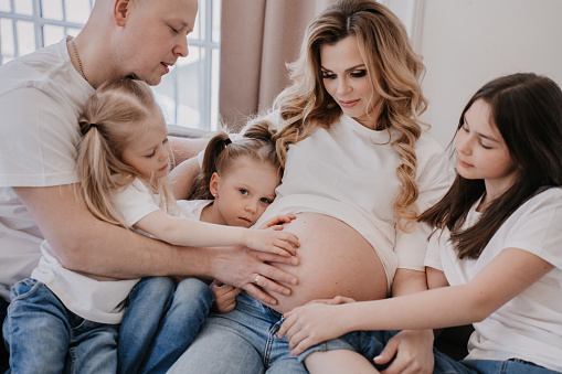 Family dressed in white t-shirts and blue jeans gathered around pregnant mother, touching her belly sitting on sofa at home. Indoor shoot of a pregnant swedish woman with loving, carrying family.