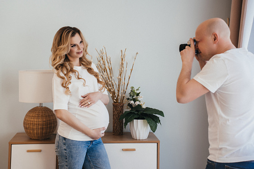 Mid aged bald man dressed in white t-shirt and blue jeans making  photos of his beautiful pregnant wife at home, standing close to locker, lamp and vase on it. Happy pregnant woman posing at home.