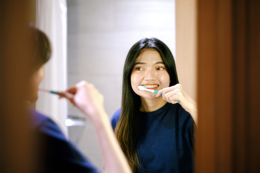 Beautifuk young adult woman brushing her teeth looking at mirror