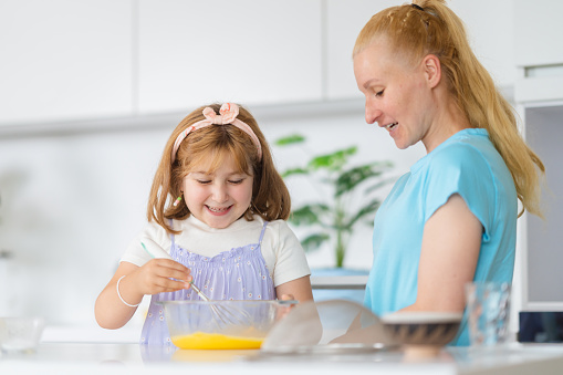 A small girl is enjoying making a cake with her mother in the kitchen at home.
