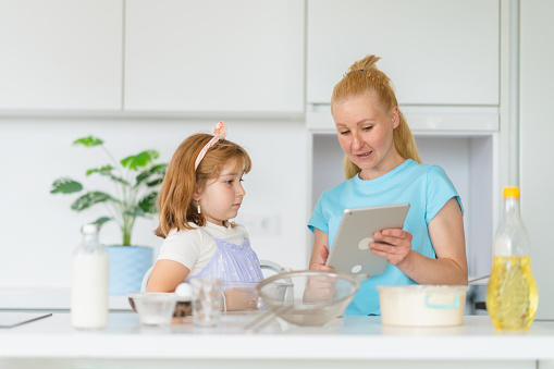 A small girl is enjoying making a cake with her mother in the kitchen at home. They are using a digital tablet to check the recipe.