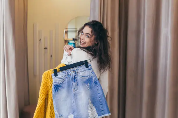 Photo of Young beautiful happy woman girl in a clothing store in the mall, holding on his shoulders, in his hands hangers with colored clothes, goes to the fitting room with a mirror to measure clothes, rejoices, smiles, chooses, looks at the camera