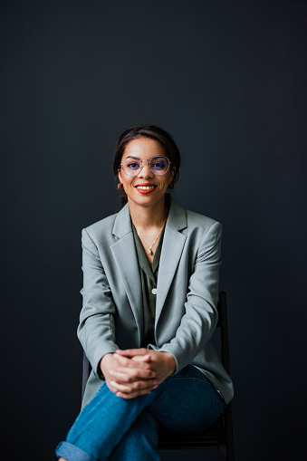 Happy businesswoman with eyeglasses looking at camera while sitting on a chair in front of the black wall.