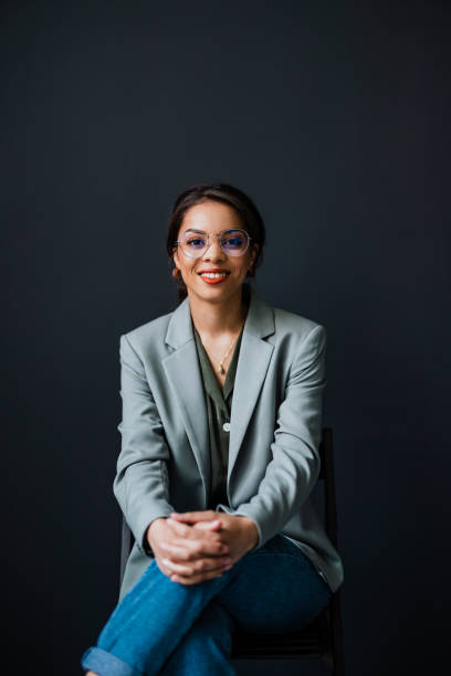 Portrait Of Smiling Business Woman Sitting With Crossed Legs At Her Office Happy businesswoman with eyeglasses looking at camera while sitting on a chair in front of the black wall. woman sitting cross legged in chair stock pictures, royalty-free photos & images
