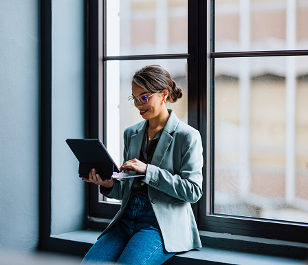 Smiling businesswoman with eyeglasses leaning on a window and typing business report on a digital tablet keyboard.