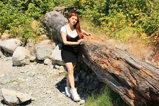 A Mexican woman on a beach with rocks and a washed up log. She is wearing a white short sleeved shirt, black short skirt and white shoes.
