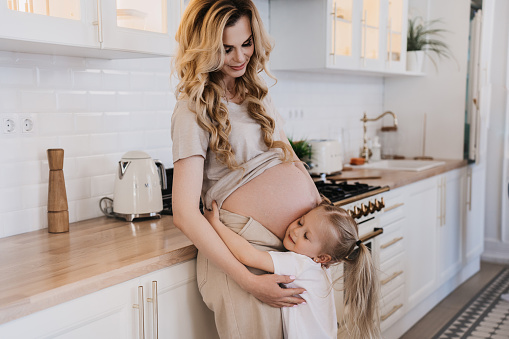 Indoor shoot of a little girl in white t-shirt hugging her pregnant mothers belly, putting her ear to hear her brother or sister. Girl embracing mom at the kitchen. Pregnancy and maternity concept.