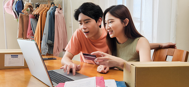 young asian couple small business owners packing using laptop together viewing their sales performance by table with many parcel at home - woman takes credit card to receive income transfer