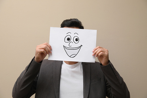 Man hiding emotions using card with drawn smiling face on beige background