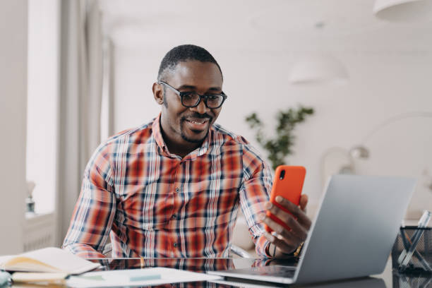 istock Happy afro man has video call on phone. Guy stays home on quarantine and working remotely on laptop. 1407723616