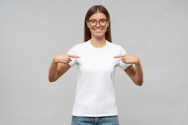 Photo of Smiling woman pointing at her blank white t-shirt with both index fingers