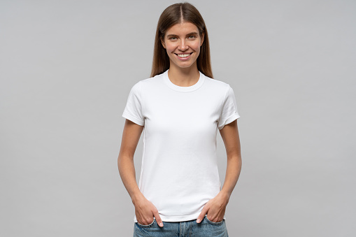 Young woman in casual white t-shirt standing with hands in pockets, isolated on gray background