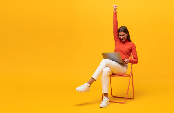 Excited young woman sitting on chair with laptop, shouting yes as winner, isolated on yellow Excited young woman sitting on chair with laptop, shouting yes as winner, isolated on yellow background with copy space chairperson stock pictures, royalty-free photos & images