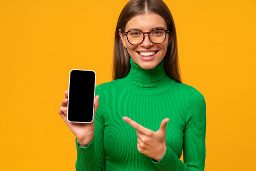 Happy young woman pointing to black phone screen with copy space, isolated on yellow background