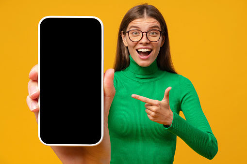 Excited shocked young woman pointing finger to blank phone screen with copy space for app, isolated on yellow background