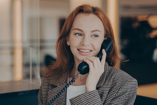 Portrait of satisfied female office worker with red hair calling business partner or client at work, using black landline phone and smiling, office environment in blurred background