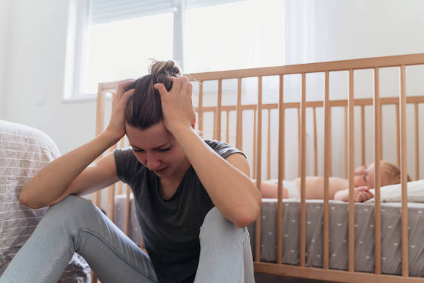 Mother holding head in hands suffering postnatal depression sitting next to baby sleeping in bed