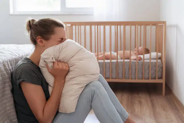 Depressed mother screaming with anger and desperation in pillow to overcome negative emotions, suffering postnatal depression because of lack of help and support with childcare routine