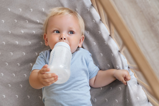 Upper view of adorable baby boy holding bottle with formula lying in crib, enjoying milk or tasty healthy nutritious baby formula. Bottle-feeding concept. Childcare