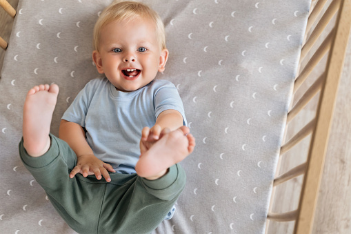 Upper view of sweet cute lovely adorable innocent blond baby with blue eyes resting in bed having fun touching his foot putting legs up. Happy carefree childhood