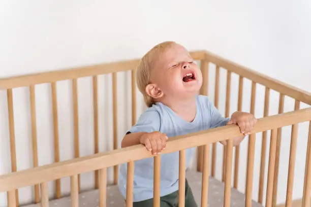 Photo of Portrait of upset crying baby standing in crib getting hysterical seeking attention of parents