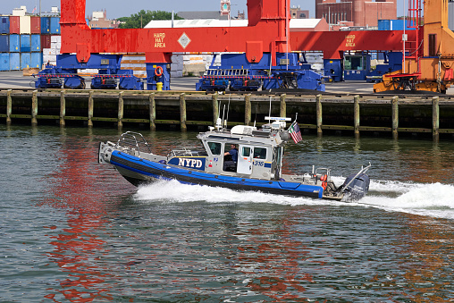 Brooklyn, NY - June 13, 2022: A NYC Police Dept. inflatable boat patrols New York Harbor. Behind is the track and wheels of gantry cranes at Red Hook Container Terminal in Red Hook, Brooklyn, NYC. The boat is a rigid hull inflatable boat (RHIB) with outboard motors.