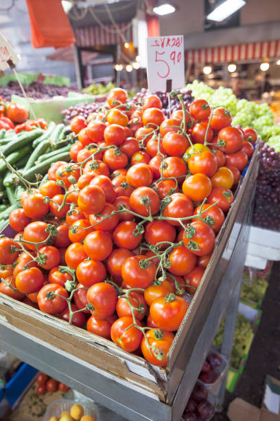Fresh, vibrant red tomatoes on the vine, with cucumbers and grapes in the background, on display at Carmel Market in Tel Aviv.