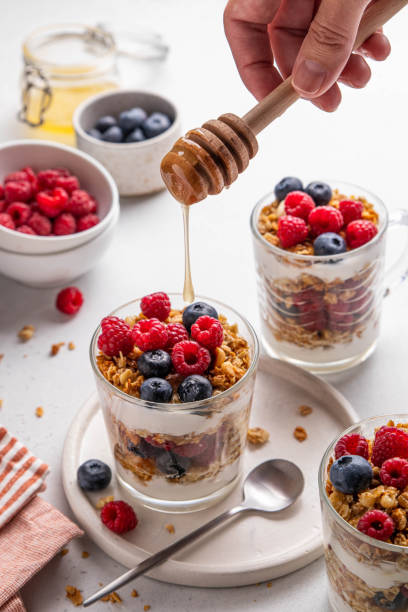 granola com iogurte em vidro servido com framboesa fresca, mirtilo e mel. muesli de aveia caseiro. processo de derramar mel na granola. mel escorre de colher de pau na mão. foco seletivo. - jar oatmeal granola glass - fotografias e filmes do acervo