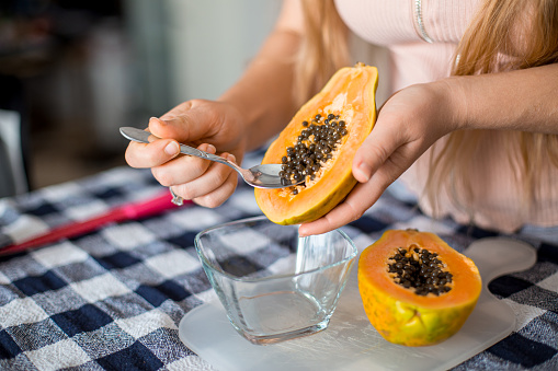 Closeup of  young woman removing seeds with tea spoon, deseeding two halves ripe fresh orange papaya fruit slices