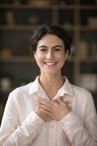 Happy grateful millennial Hispanic girl applying hands to chest in gesture of thank, gratitude. Young woman making sign of kindness, showing symbol of love, care, support. Vertical shot