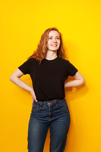 Studio portrait of an attractive 24 year old red-haired woman in a black t-shirt and jeans on a yellow background