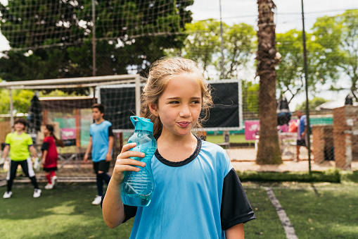 Portrait of a cute girl drinking water during a soccer training. Other kids playing soccer in the background.