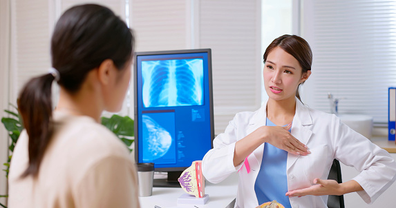 attractive young brunette ponytail female asian doctor wear white coat with pink ribbon explaining breast self examination to woman at clinic - a model on table and xray display