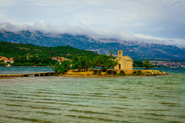Small island with some green trees and small old church on it with mountains in background and cloudy sky