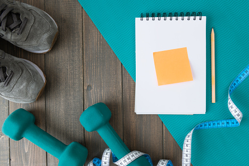 Directly above view of green dumbbell and green mat, sport shoes, notepad and yellow post-it on wooden background.