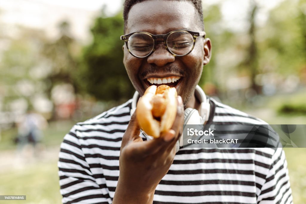 Close up portrait of young man eating hot dog outdoors Young African American man is eating a hot dog and enjoying Hot Dog Stock Photo