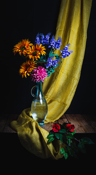 Still life of a glass vase with three roses of various colors, with several fallen flowers and wooden background.