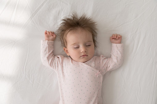 Cute baby in pink comfort bodysuit and dry diaper sleeping on bed with arms raised and eyes closed looks carefree feels peaceful, top view.  Sweet babyhood, healthy day nap for newborn growth concept