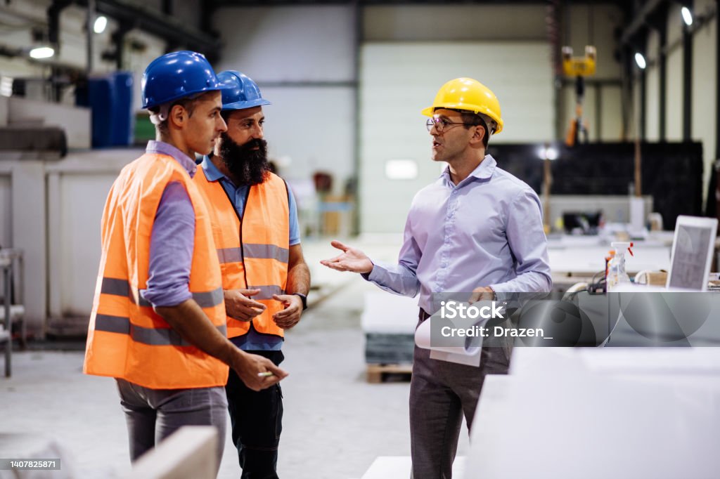 Engineers in factory hall, performing quality control. Manufacturing occupation in modern industrial hall, with automated process and robots. Metal processing industry and warehouse. Employees in factory working together Occupational Safety And Health Stock Photo