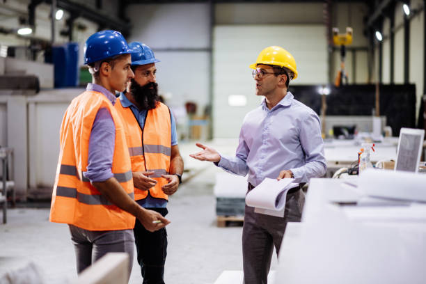 Engineers in factory hall, performing quality control. Manufacturing occupation in modern industrial hall, with automated process and robots. Metal processing industry and warehouse. Employees in factory working together health and safety stock pictures, royalty-free photos & images