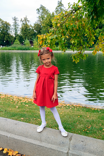 little girl with red hairpins and dress stands near pond outdoor in autumn