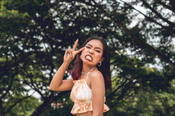 A young teenage woman makes a V-sign while grinning. Teasing and having fun while posing. Outdoor park scene.